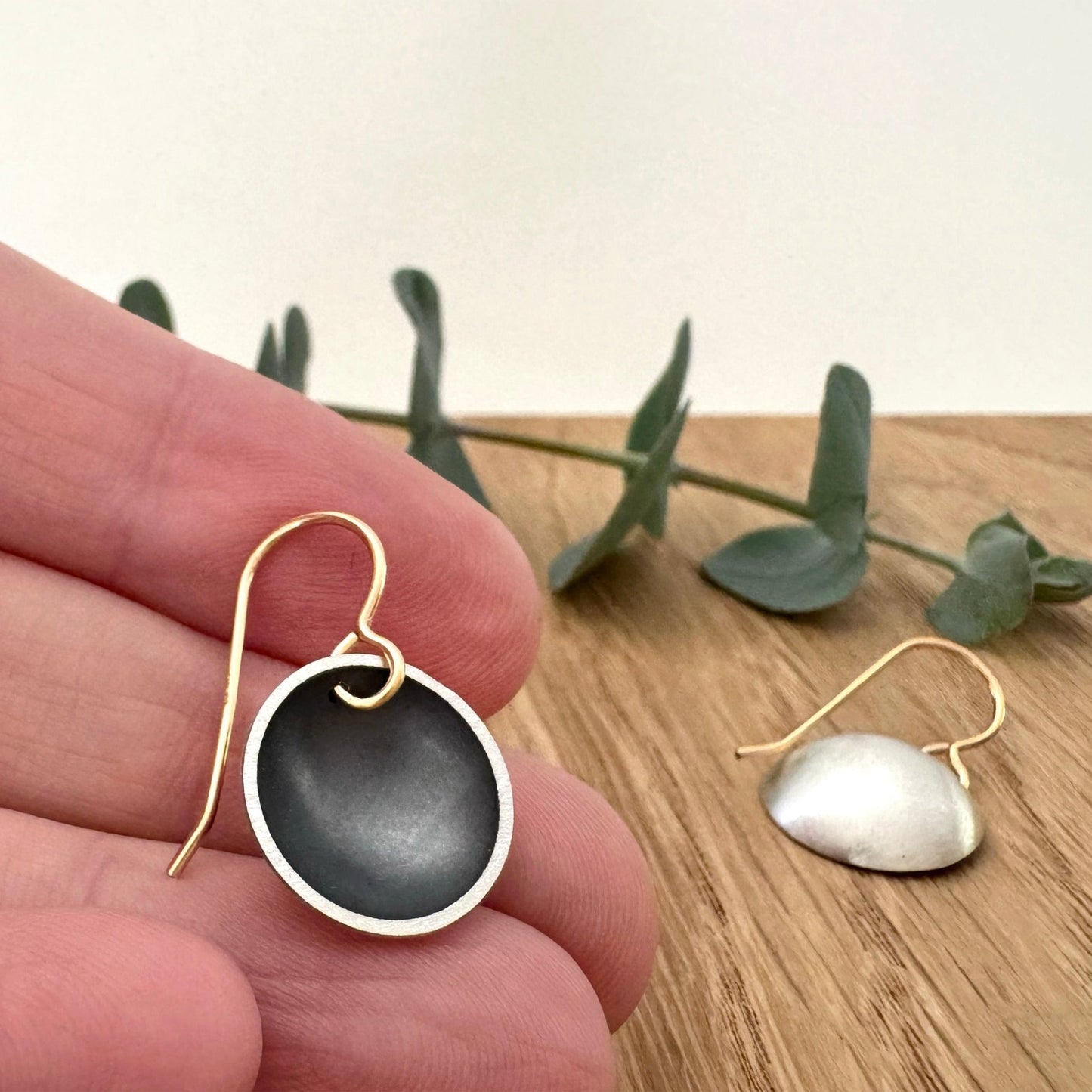 A hand holding one of a pair of oxidised sterling silver disc dangle earrings with 14/20Y gold filled ear wires, against a wooden background with a small sprig of eucalyptus leaves.