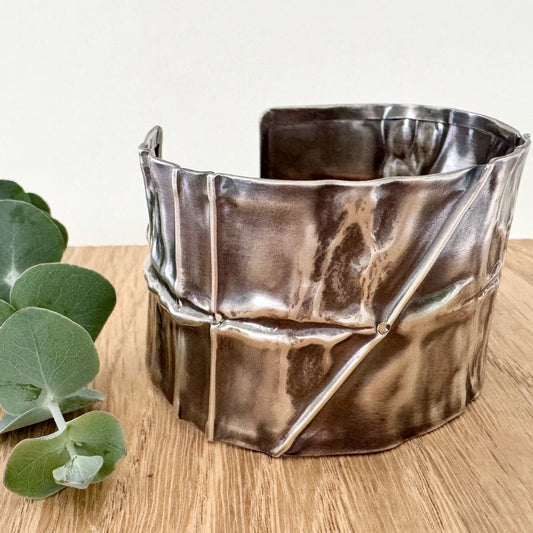 A wide, oxidised, foldformed sterling silver cuff bracelet. The cuff is highly textured and and is displayed on a wooden surface with a small sprig of eucalyptus leaves.