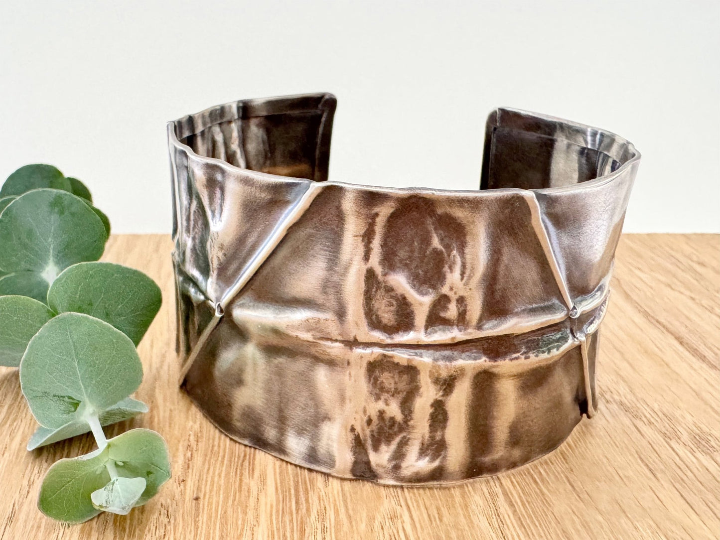 A wide, oxidised, foldformed sterling silver cuff bracelet. The cuff is highly textured and and is displayed on a wooden surface with a small sprig of eucalyptus leaves.