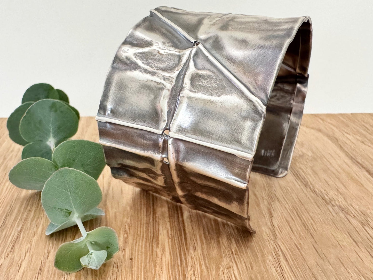 A wide, oxidised, foldformed sterling silver cuff bracelet. The cuff is highly textured and and is displayed on a wooden surface with a small sprig of eucalyptus leaves.