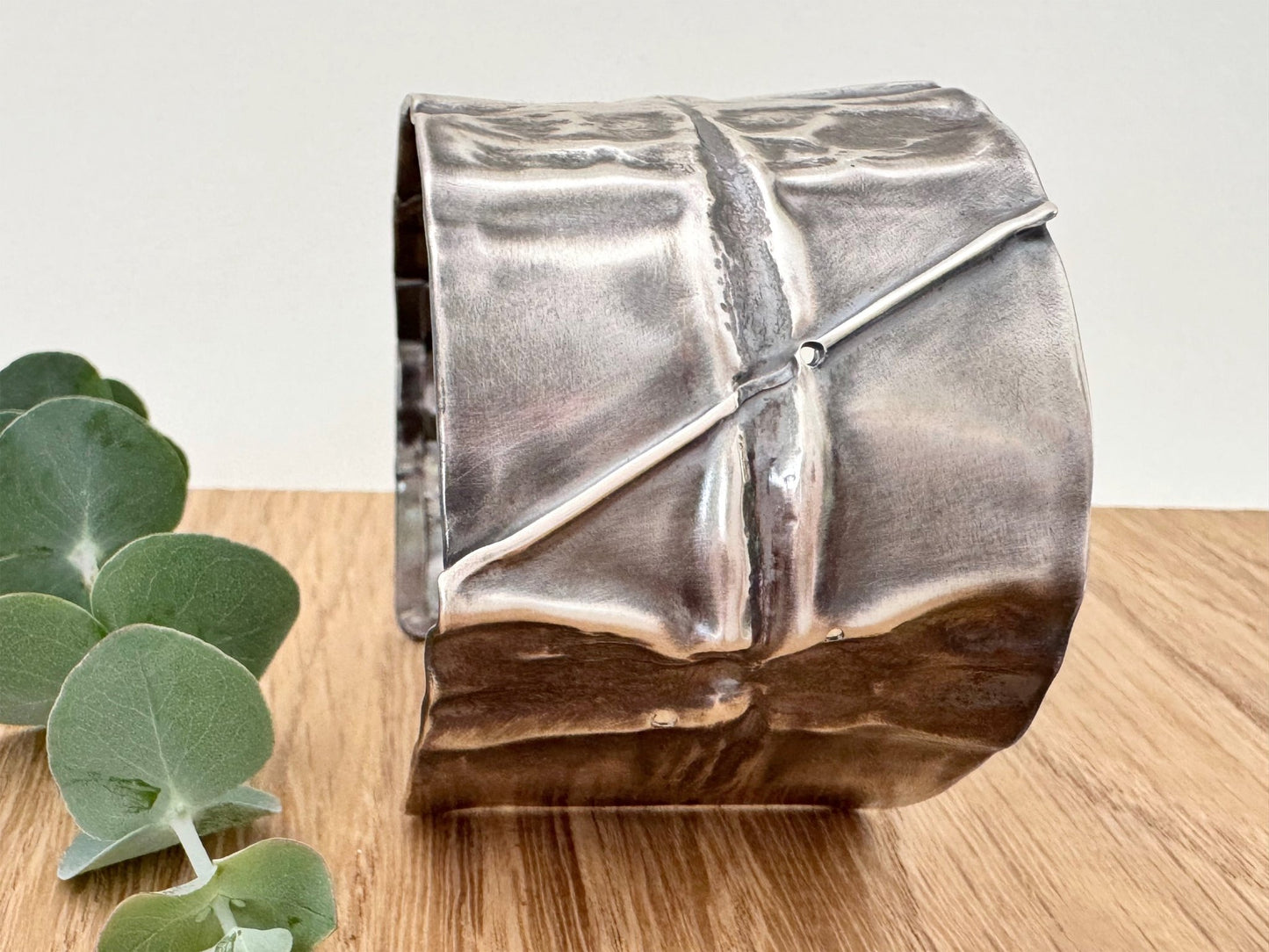A wide, oxidised, foldformed sterling silver cuff bracelet. The cuff is highly textured and and is displayed on a wooden surface with a small sprig of eucalyptus leaves.