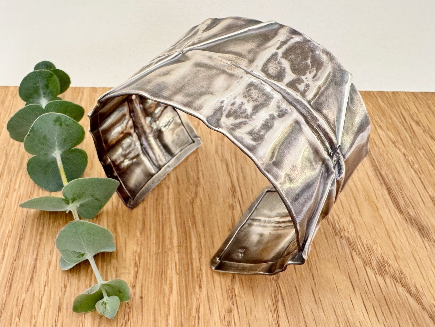 A wide, oxidised, foldformed sterling silver cuff bracelet. The cuff is highly textured and and is displayed on a wooden surface with a small sprig of eucalyptus leaves.