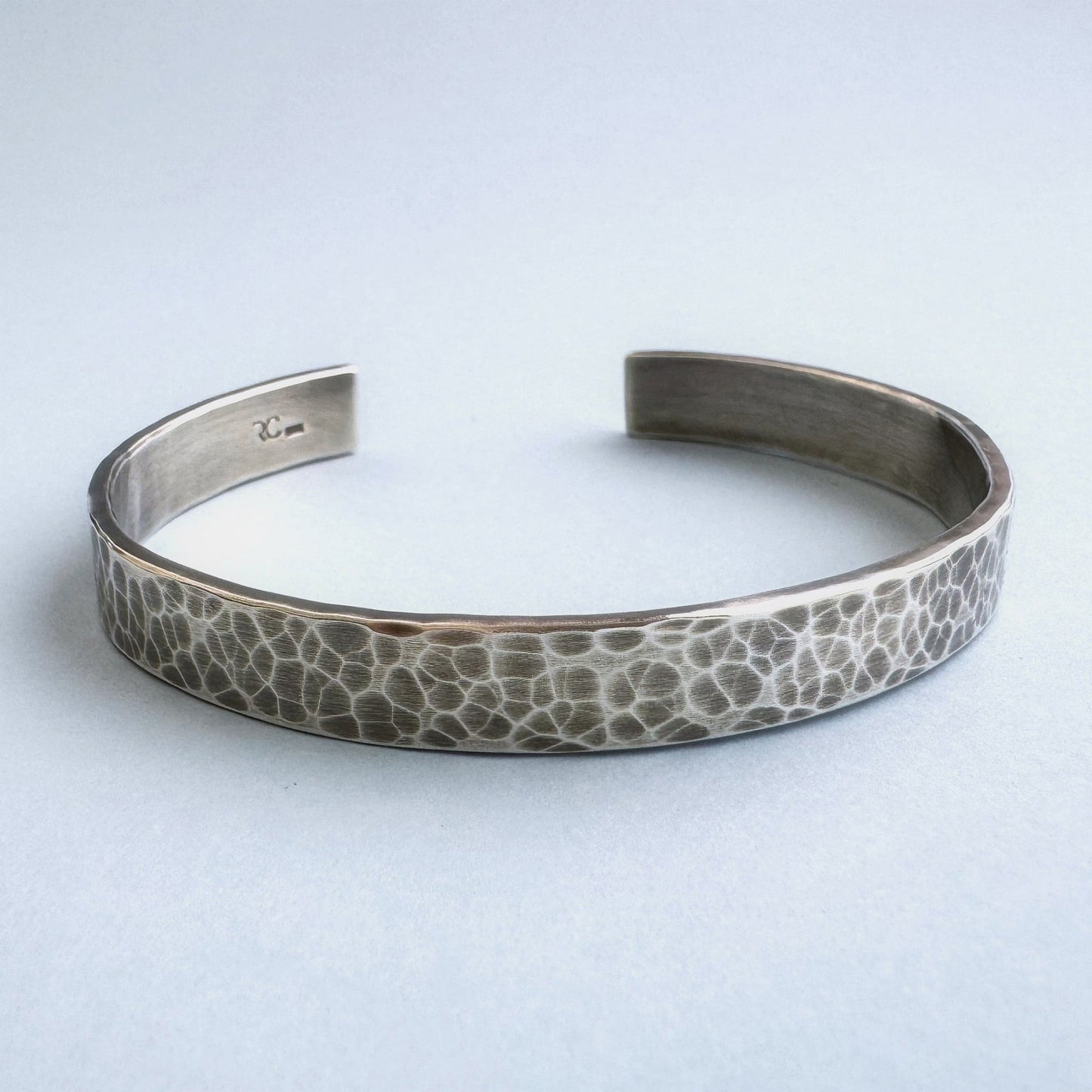 A heavy, hammered/beaten sterling silver cuff bracelet with a blackened finish is lying on a pale blue background.