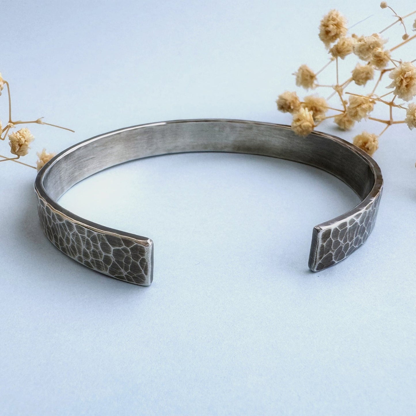 A heavy, hammered/beaten sterling silver cuff bracelet with a blackened finish is lying on a pale blue background. There are cream coloured dried flowers behind it.