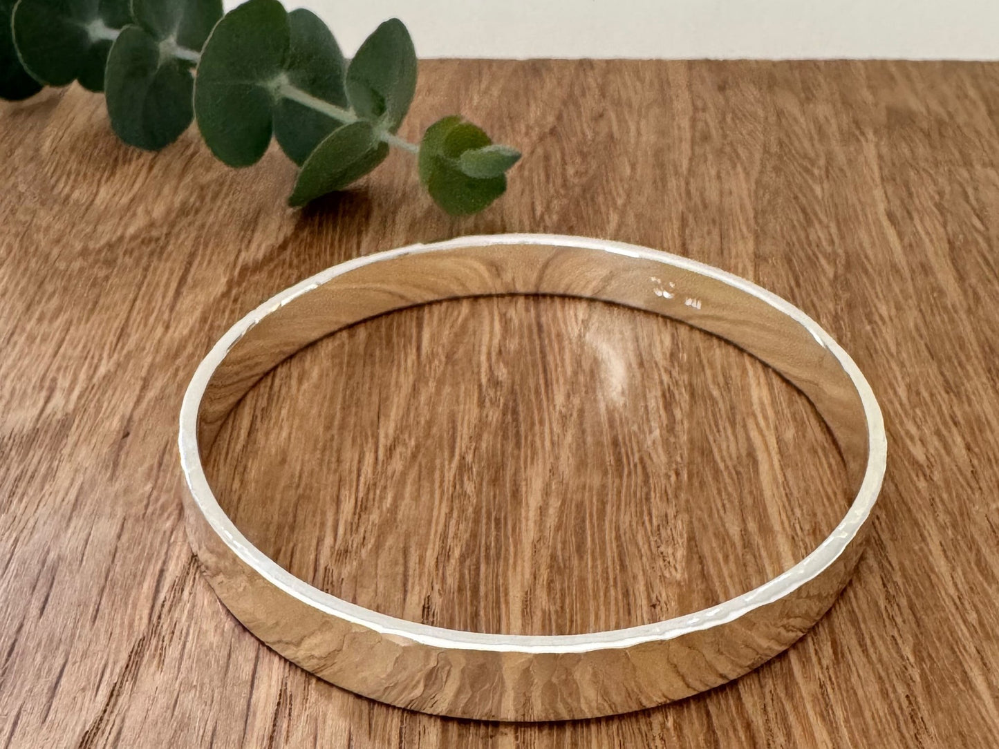 Hammered sterling silver bangle on a wooden surface with a small sprig of eucalyptus leaves