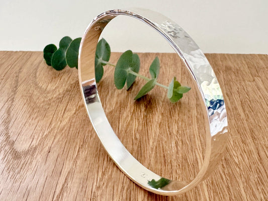 Hammered sterling silver bangle on a wooden surface with a small sprig of eucalyptus leaves
