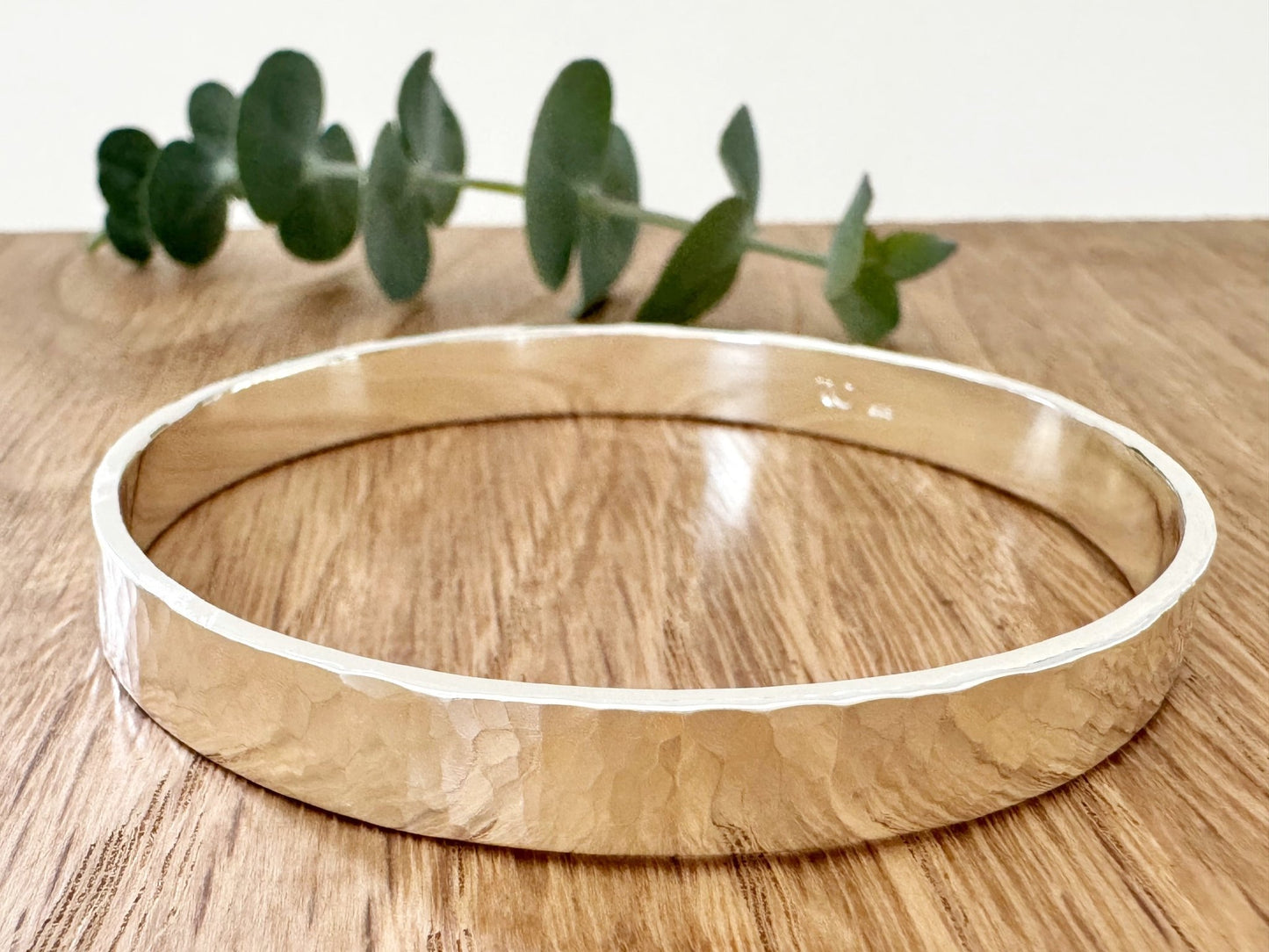 Hammered sterling silver bangle on a wooden surface with a small sprig of eucalyptus leaves