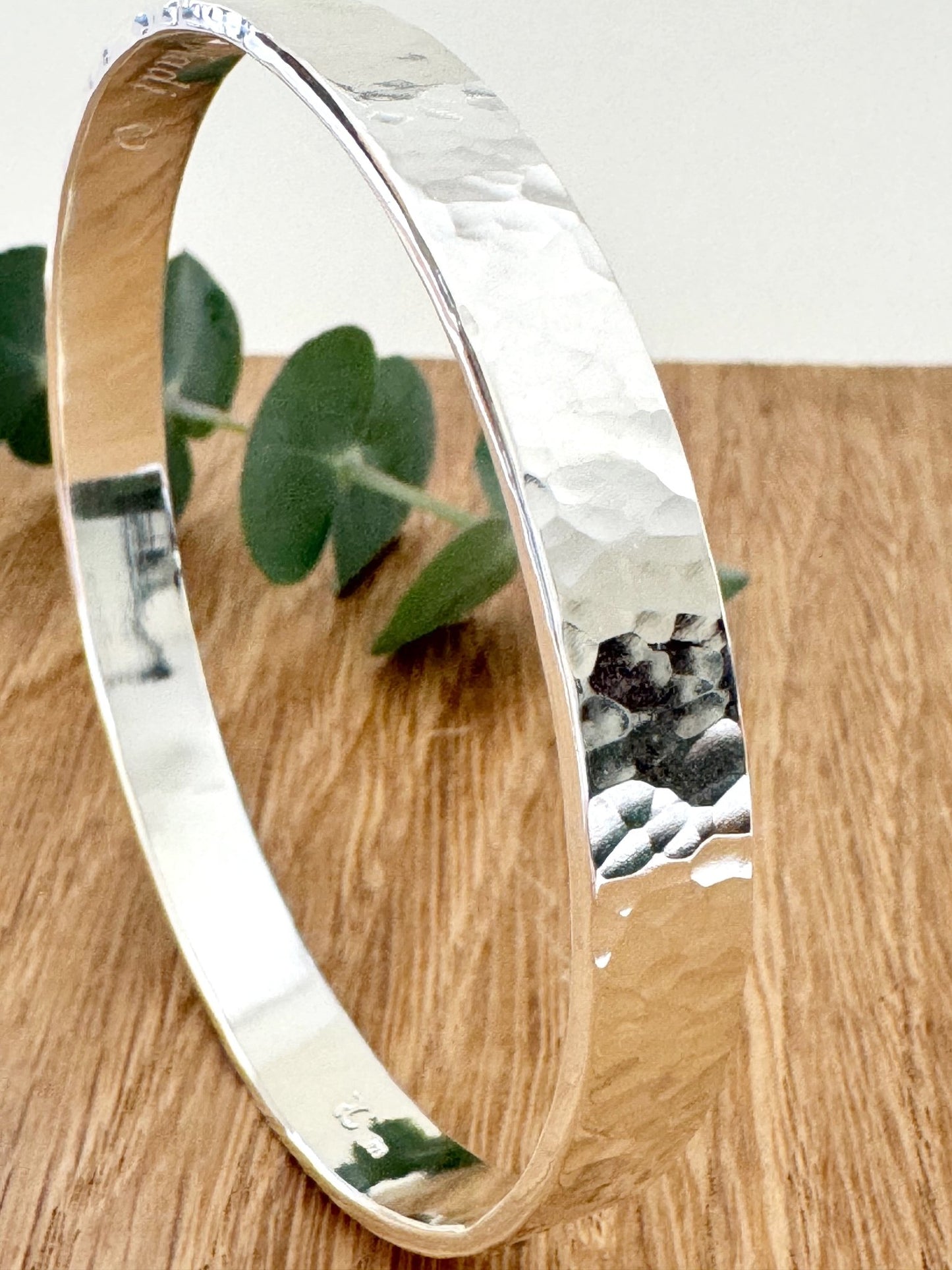 Hammered sterling silver bangle on a wooden surface with a small sprig of eucalyptus leaves