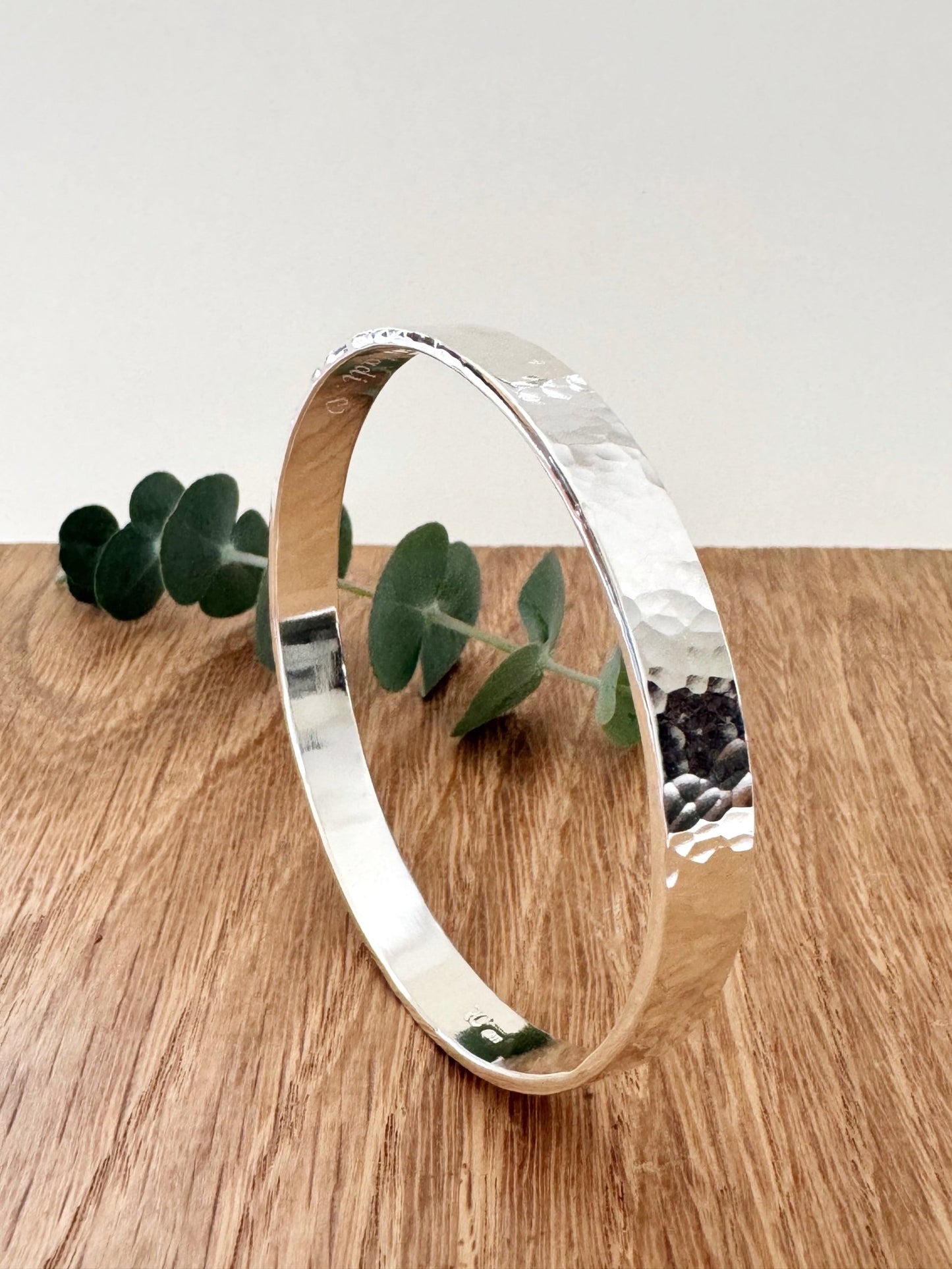 Hammered sterling silver bangle on a wooden surface with a small sprig of eucalyptus leaves
