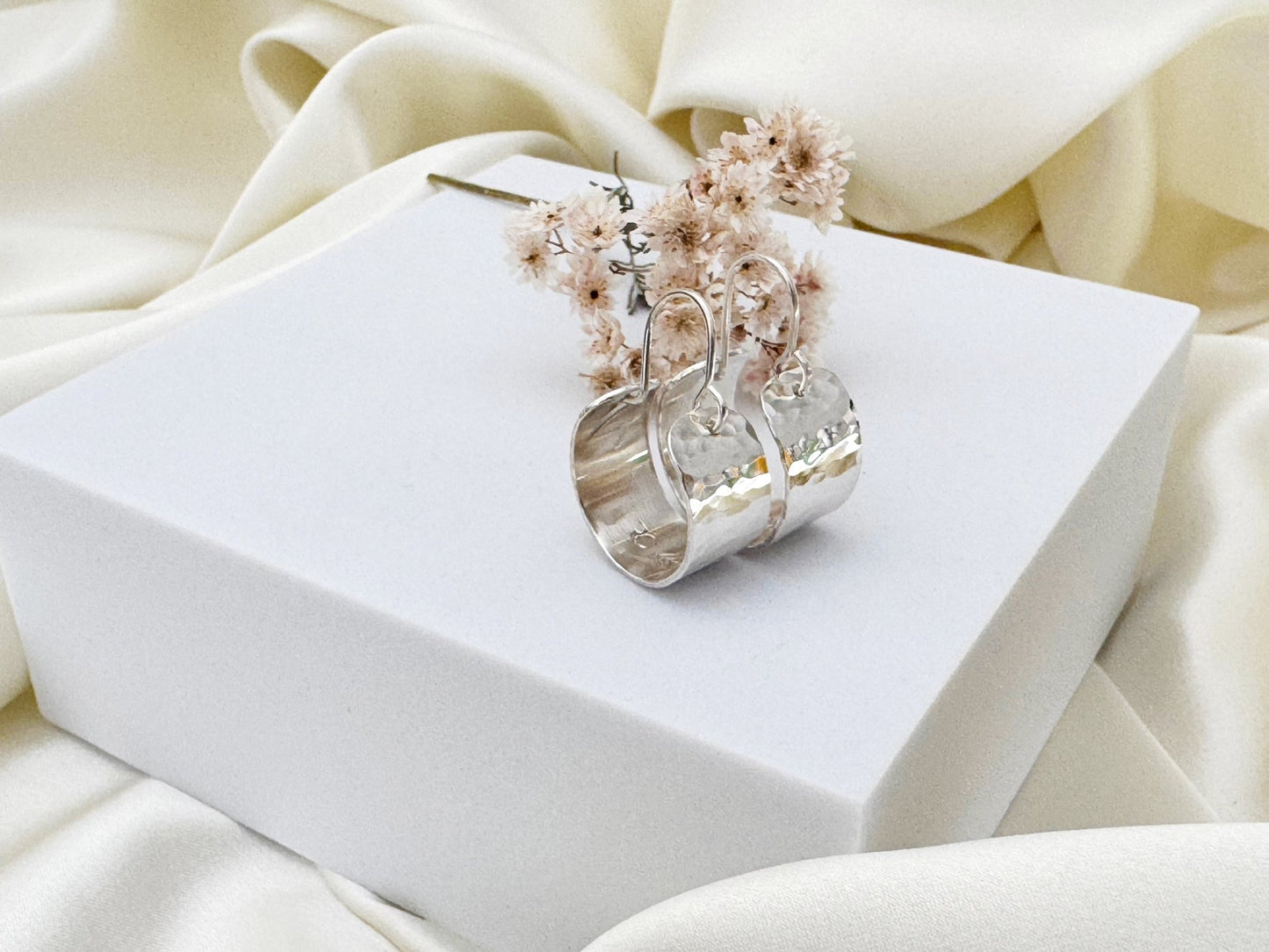 Silver hoop earrings with hammered texture on a white box against a beige fabric background