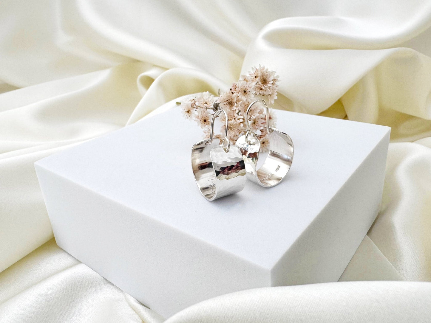 Silver hoop earrings with a hammered texture on a white box against a soft white fabric background