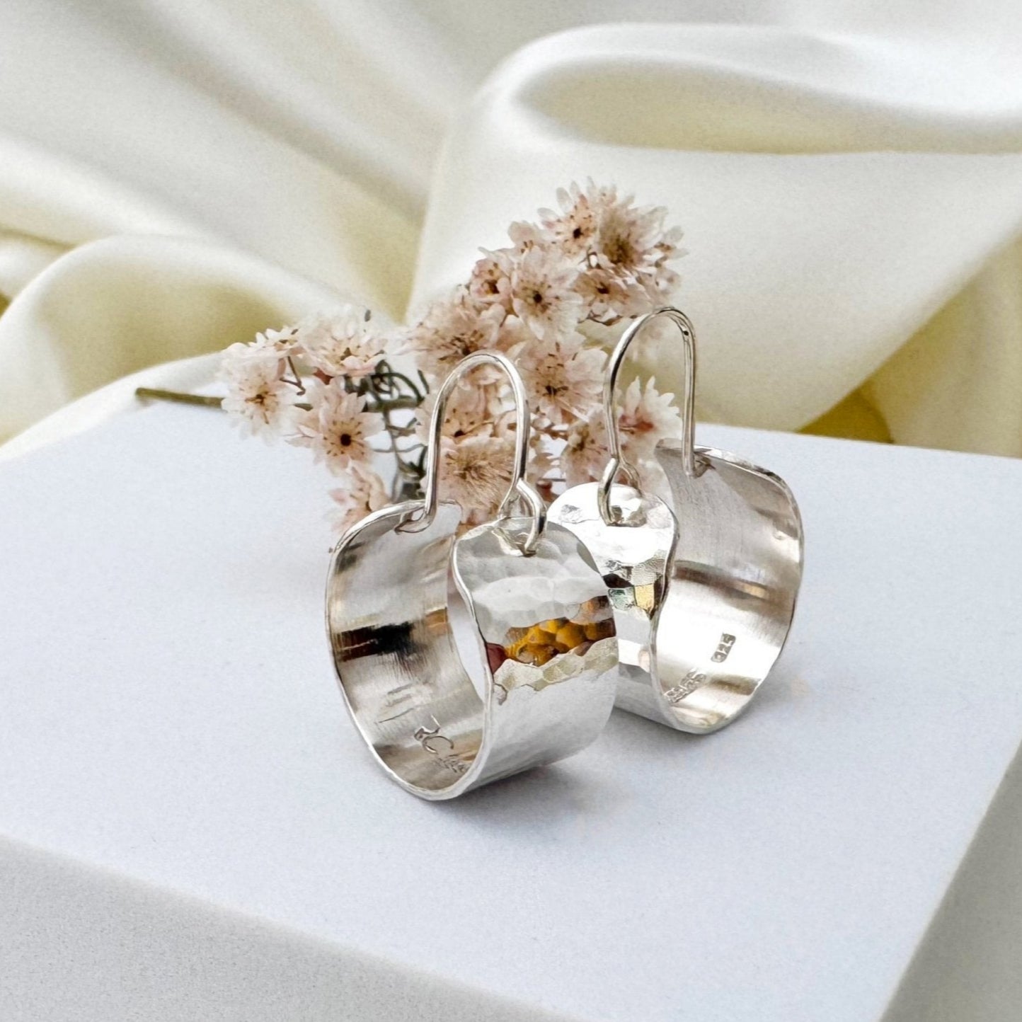 Silver hoop earrings with hammered texture on a white surface in front of a cream satin background