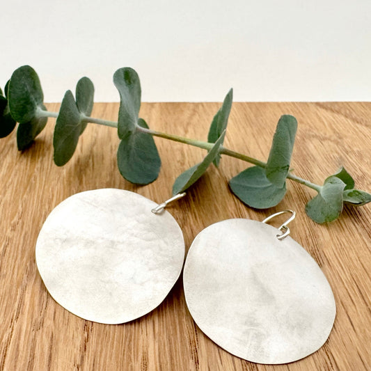 A pair of large, hammered, brush sterling silver disc earrings rests on a wooden surface next to a small sprig eucalyptus leaves.