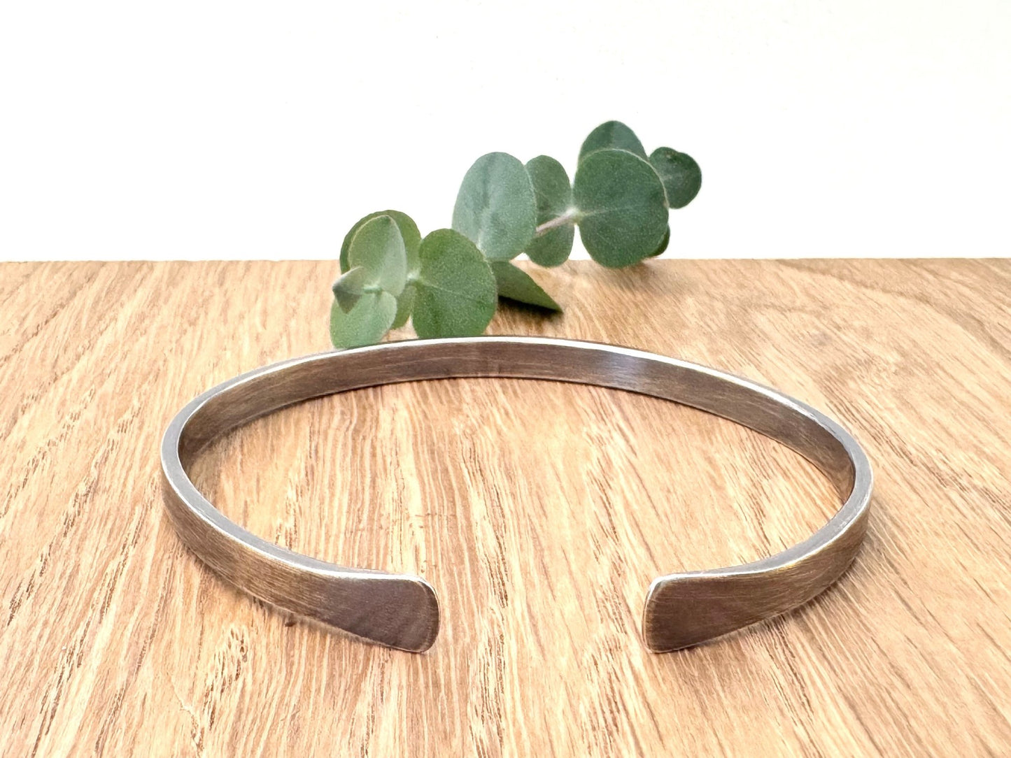 An oxidised sterling silver bracelet with flared ends on a wooden surface with eucalyptus leaves in the background.
