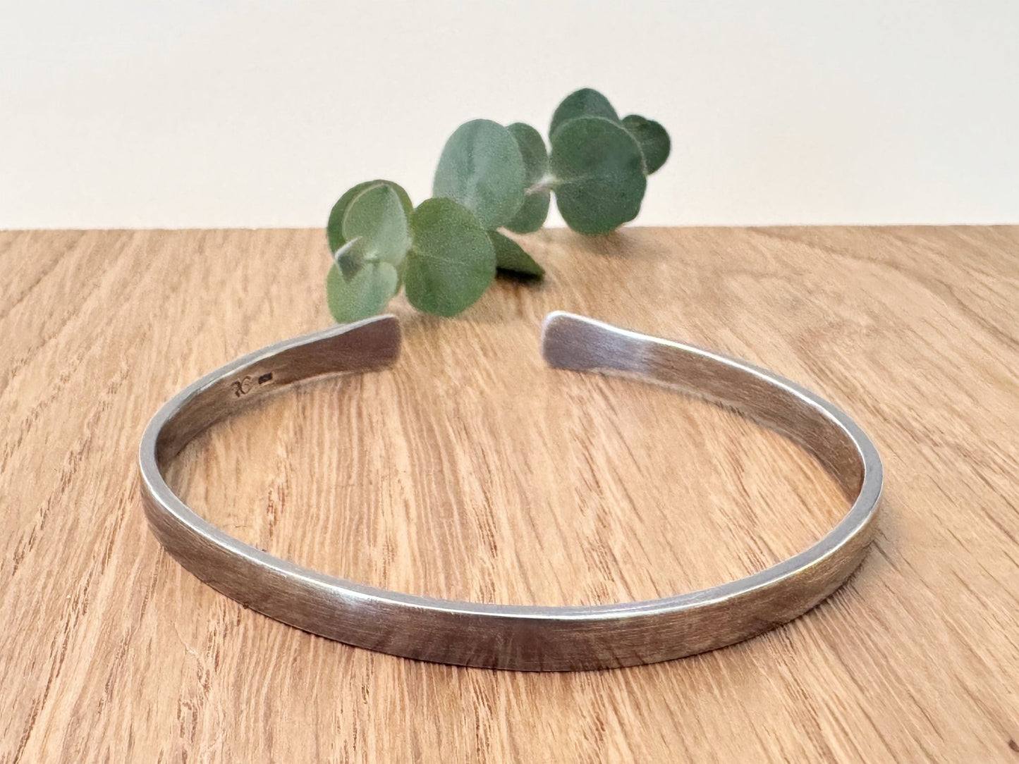 An oxidised sterling silver bracelet with flared ends on a wooden surface with eucalyptus leaves in the background.