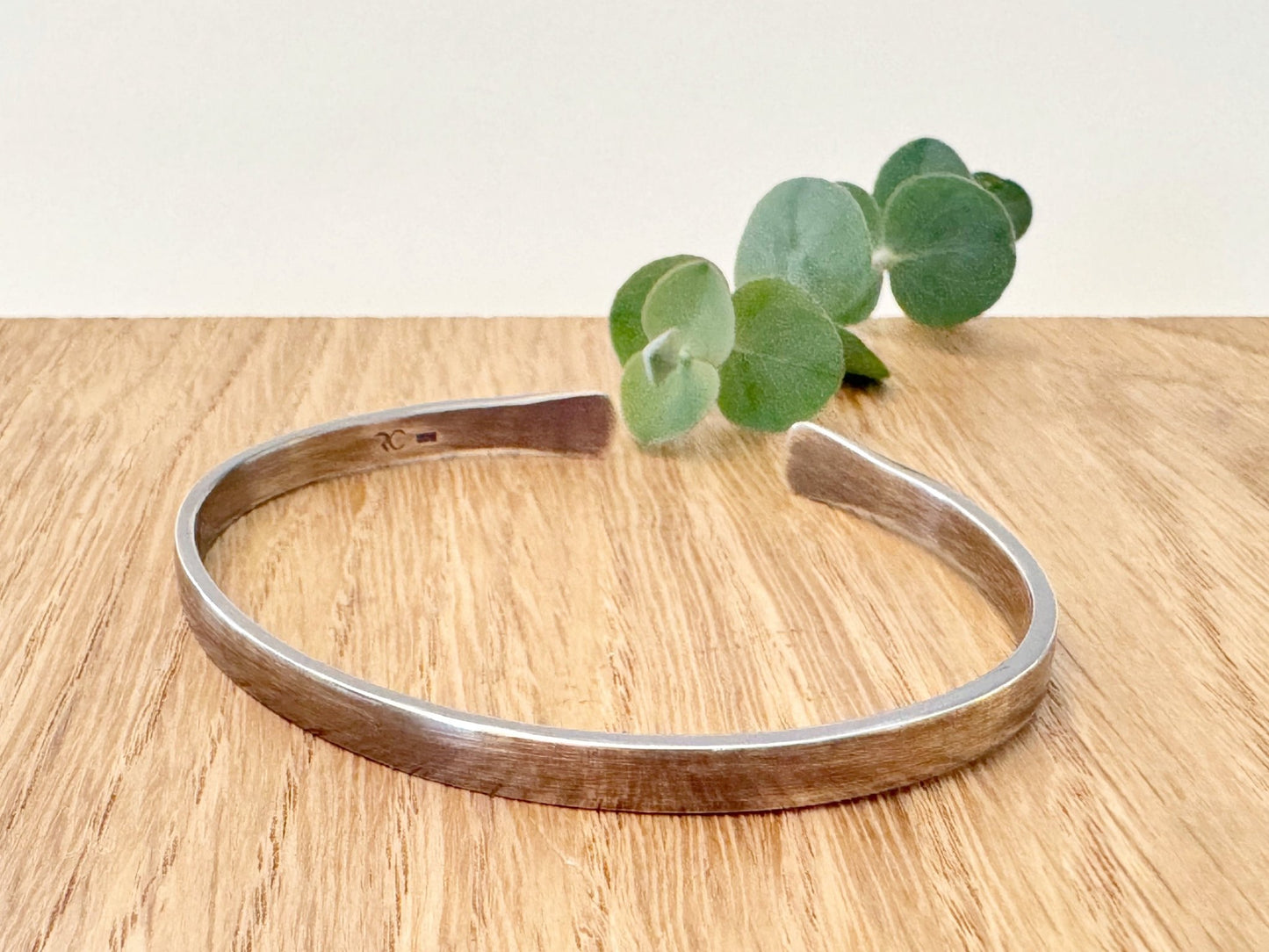 An oxidised sterling silver bracelet with flared ends on a wooden surface with eucalyptus leaves in the background.