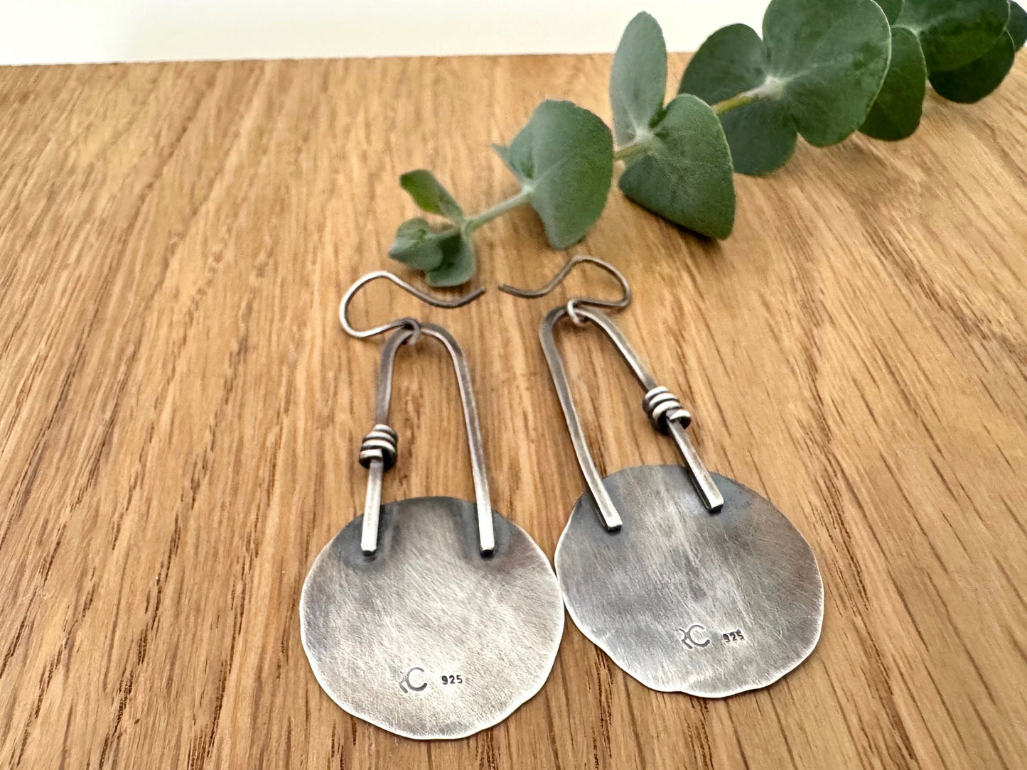 Pair of handmade oxidised sterling silver statement earrings on a wooden surface with a small sprig of eucalyptus leaves.