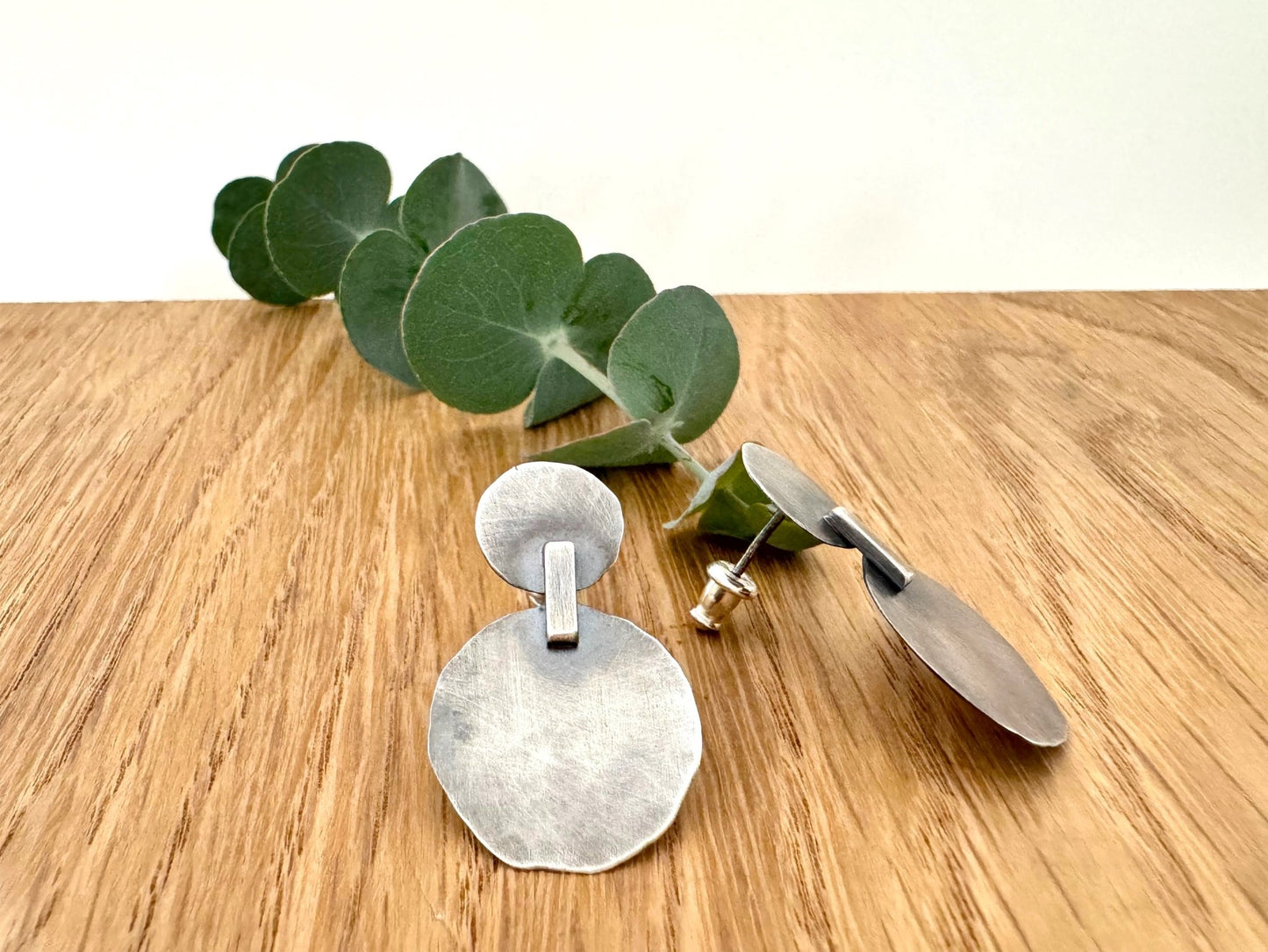 Silver earrings on a wooden surface with a small sprig of eucalyptus leaves