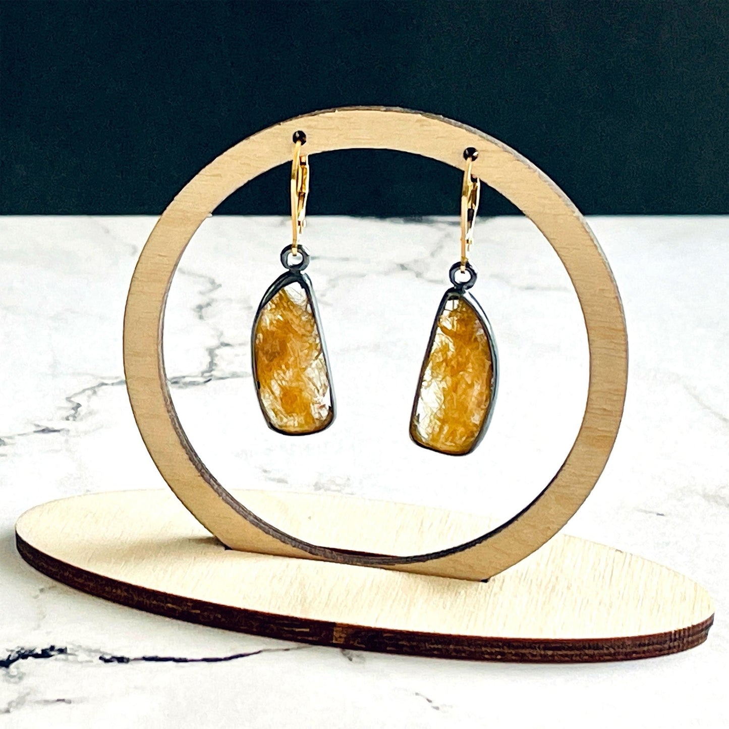 A pair of handmade sterling silver drop earrings featuring gold rutile quartz gemstones. The sterling silver bezel settings have been heavily oxidised and the earwires are 14k gold fill lever backs. The earrings are hanging from a wooden stand, which is on a white marble base with a black background.