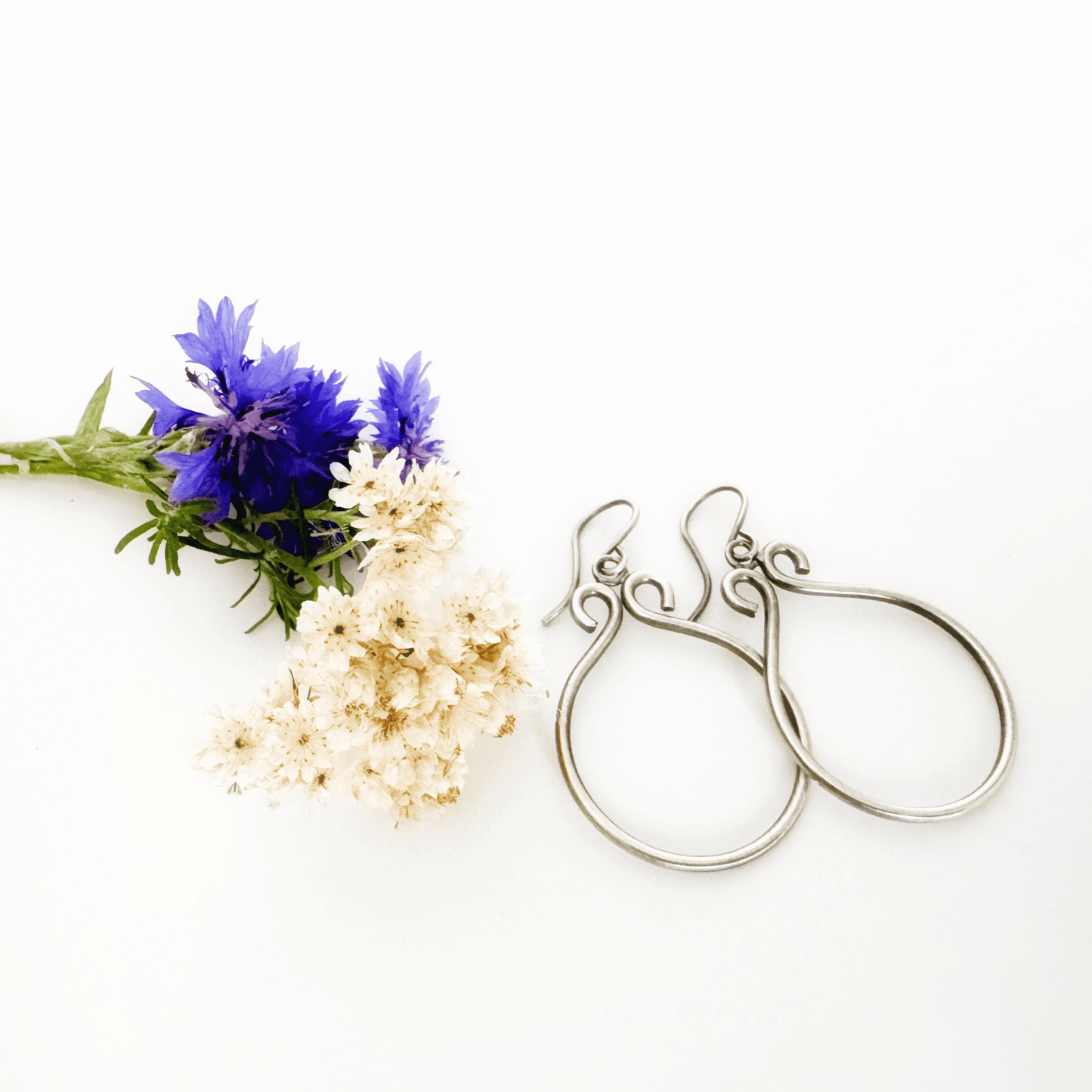 A set of sterling silver hoop earrings on a white background next to cream and blue flowers. The hoops have been oxidised.