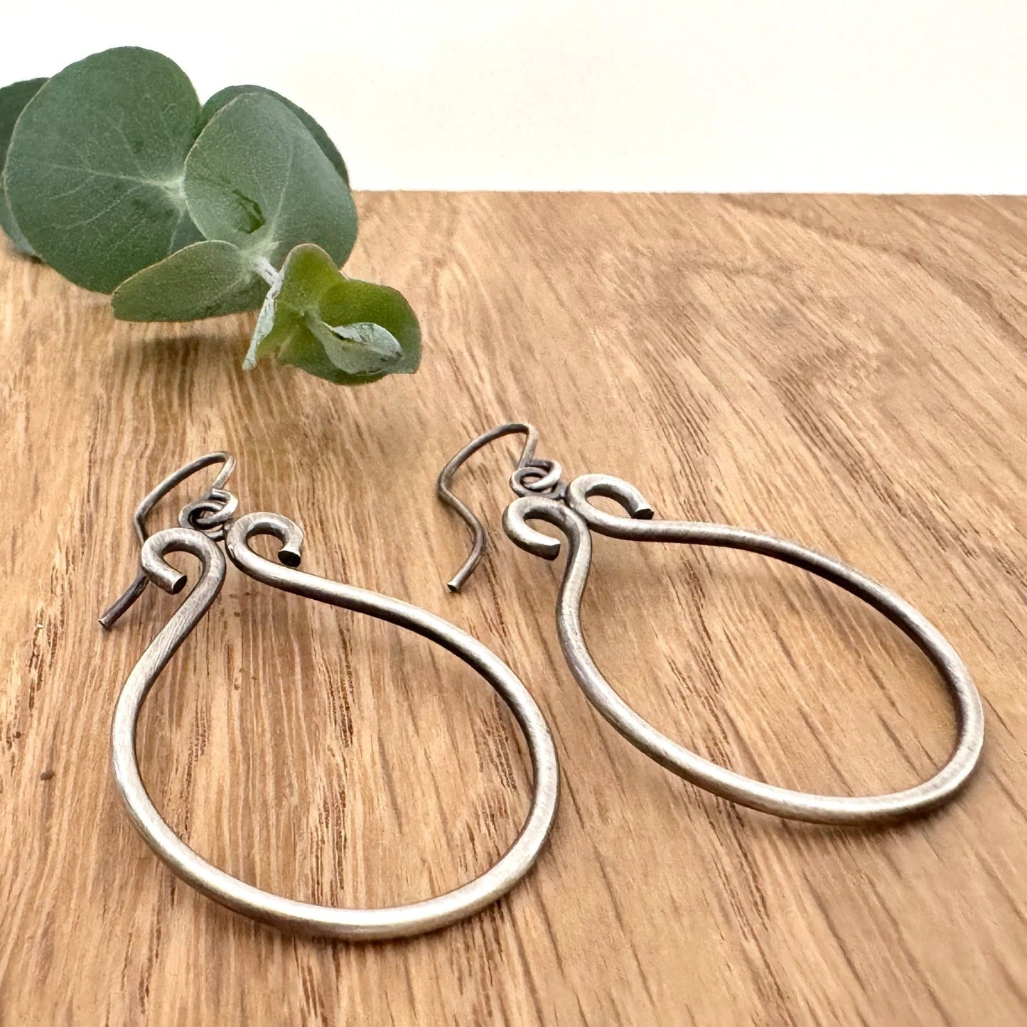 Oxidised sterling silver hoop earrings on a wooden surface with a small sprig of eucalyptus leaves.