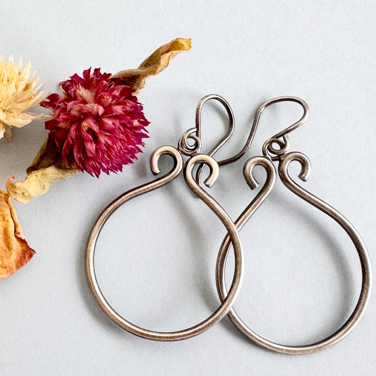 Set of handmade sterling silver hoops on a light grey background with cream, yellow and pink dried flowers. The hoops are lightly oxidised and have loops at the top.