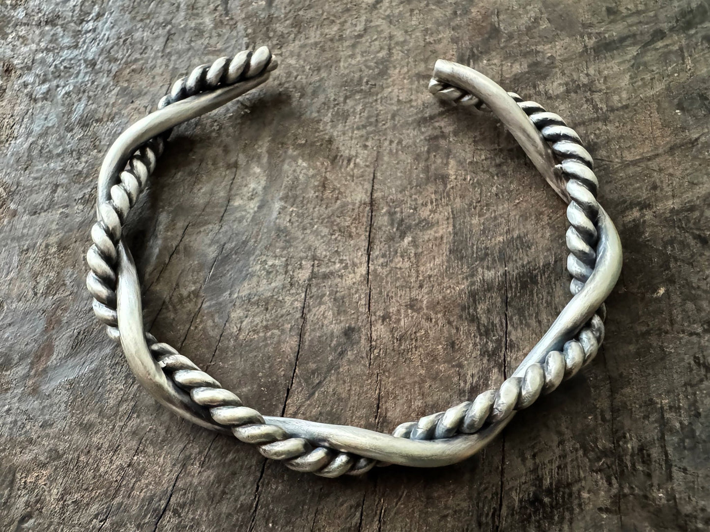 A sterling silver cuff bracelet on a wooden background. The cuff bracelet is comprised of two strands of silver, loosely twisted together. One is round and smooth, the other is very tightly twisted. It has been oxidised.