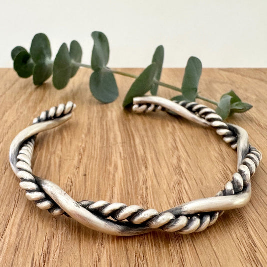 A sterling silver cuff bracelet on a wooden background with a small sprig of eucalyptus leaves. The cuff bracelet is comprised of two strands of silver, loosely twisted together. One is round and smooth, the other is very tightly twisted. It has been oxidised.