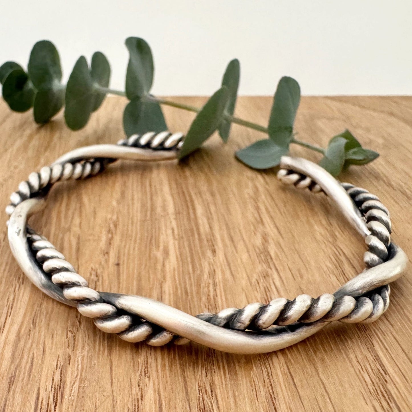 A sterling silver cuff bracelet on a wooden background with a small sprig of eucalyptus leaves. The cuff bracelet is comprised of two strands of silver, loosely twisted together. One is round and smooth, the other is very tightly twisted. It has been oxidised.