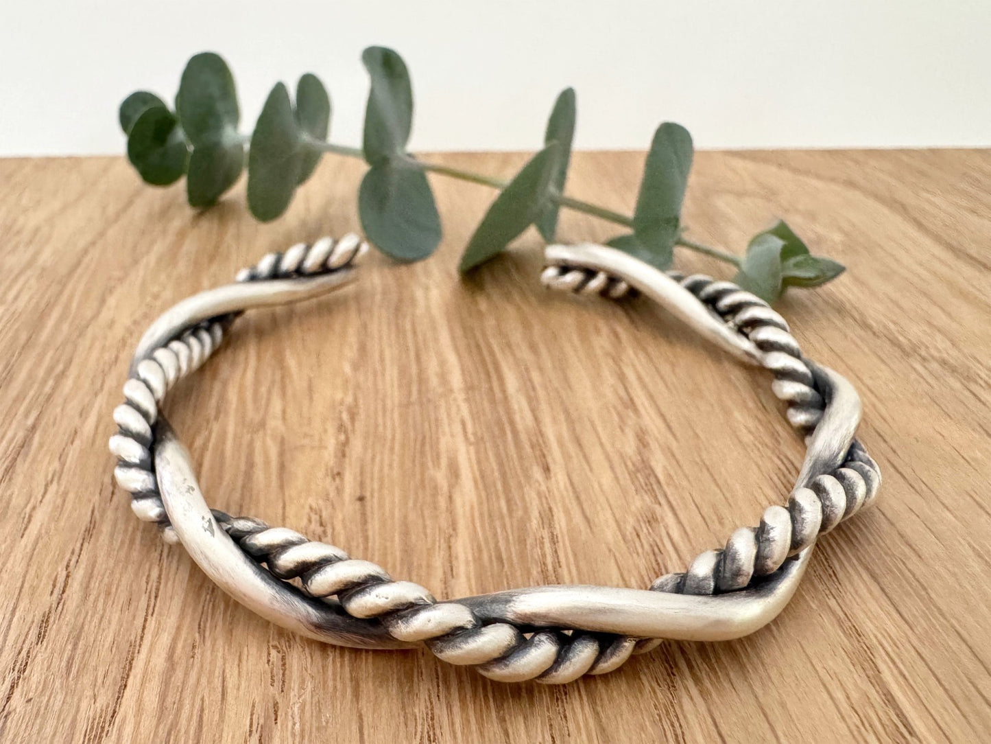 A sterling silver cuff bracelet on a wooden background with a small sprig of eucalyptus leaves. The cuff bracelet is comprised of two strands of silver, loosely twisted together. One is round and smooth, the other is very tightly twisted. It has been oxidised.