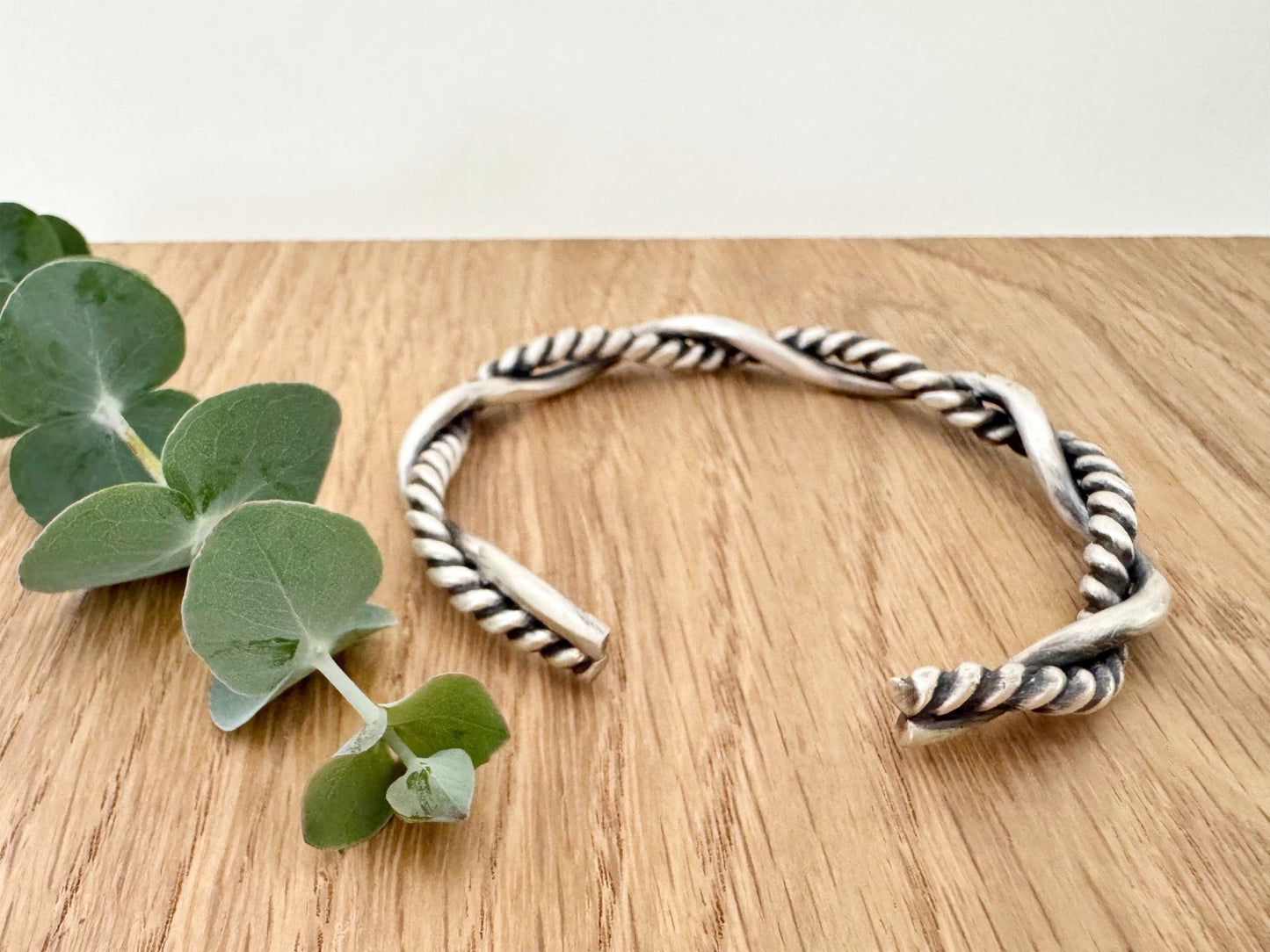 A sterling silver cuff bracelet on a wooden background with a small sprig of eucalyptus leaves. The cuff bracelet is comprised of two strands of silver, loosely twisted together. One is round and smooth, the other is very tightly twisted. It has been oxidised.