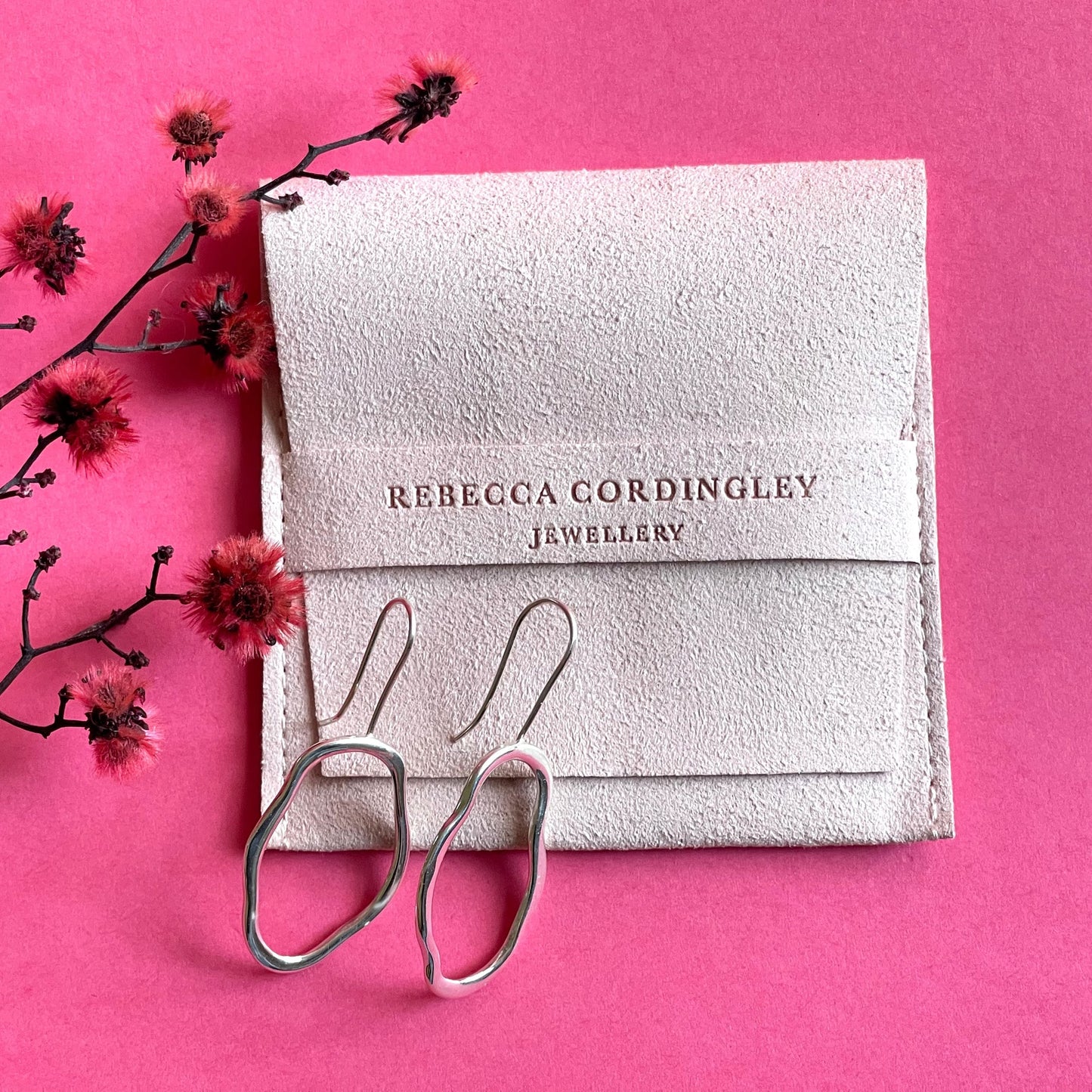 A pair of unique, abstract shaped sterling silver earrings, sitting on an ivory coloured pouch, on a bright pink background. There is dyed red blossom next to the pouch. Handmade by Australian jeweller Rebecca Cordingley.