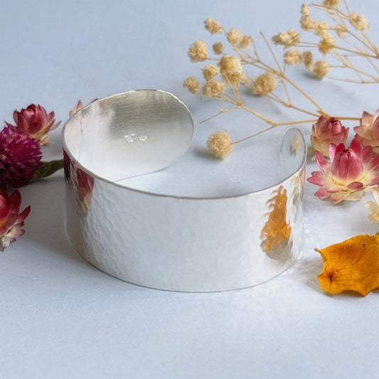 A highly polished 1 inch wide, hammered sterling silver cuff bracelet on a pale blue background, surrounded by pink, cream and yellow dried flowers - Rebecca Cordingley Jewellery