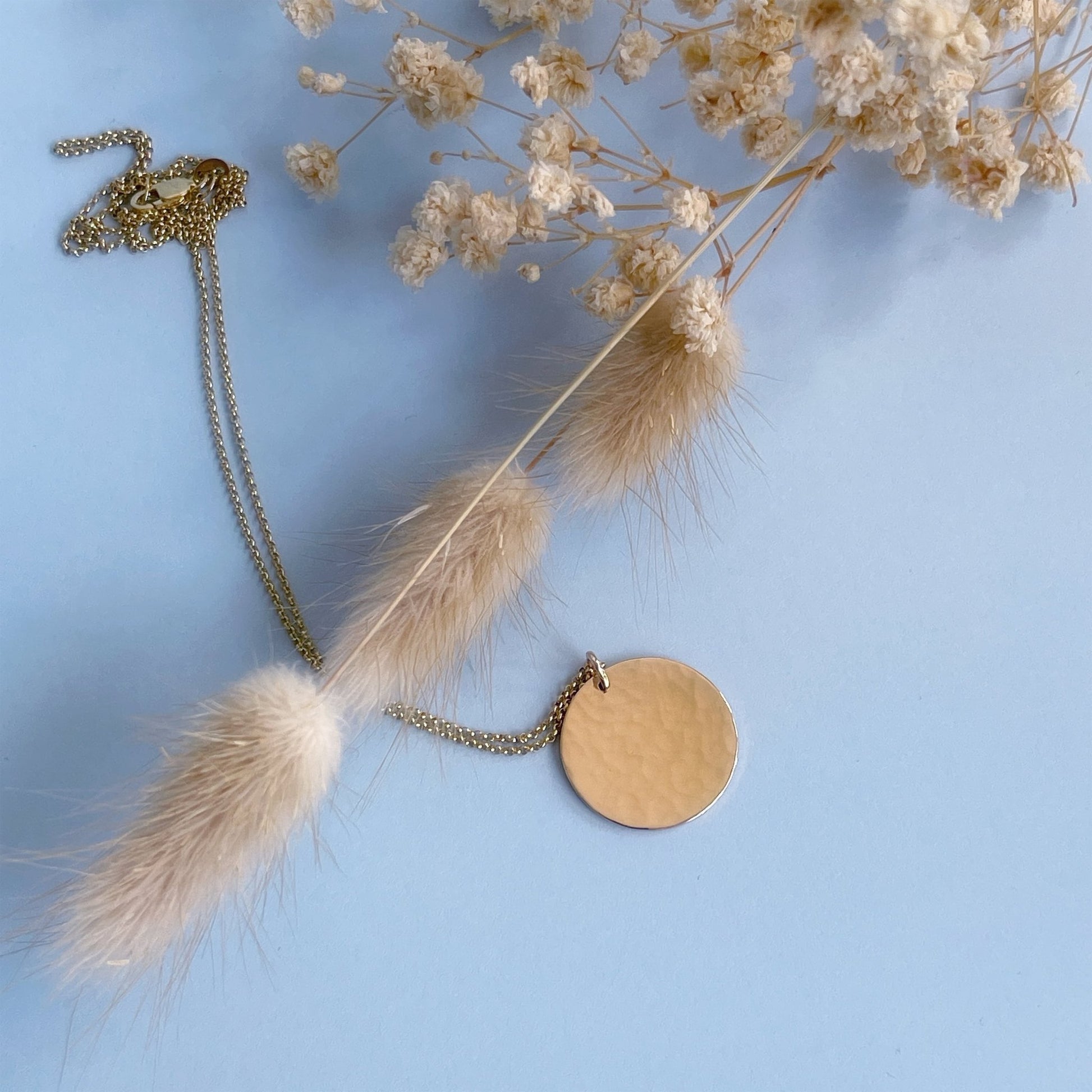 19mm diameter 14k gold coin necklace for women, on a pale blue background, with dried, cream-coloured flowers
