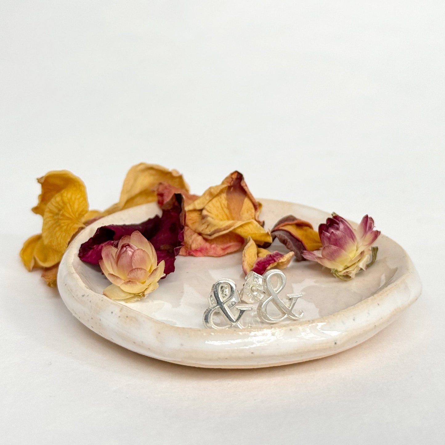 A pair of sterling silver ampersand symbol stud earrings on a small ceramic dish with dried flowers, against a cream background.