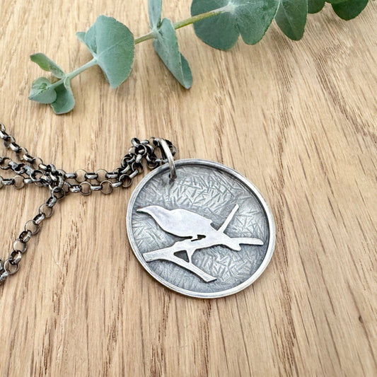 Silver necklace with a coin pendant featuring a bird design on a wooden surface with eucalyptus leaves.