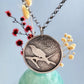 A round sterling silver pendant hangs from a rolo style chain. The pendant features a raised raven or crow sitting on the branch of a tree. The background has been hammered to represent leaves. The necklace is hanging in front of a pale green vase with cream and dark pink dried flowers, in front of a pale blue background.