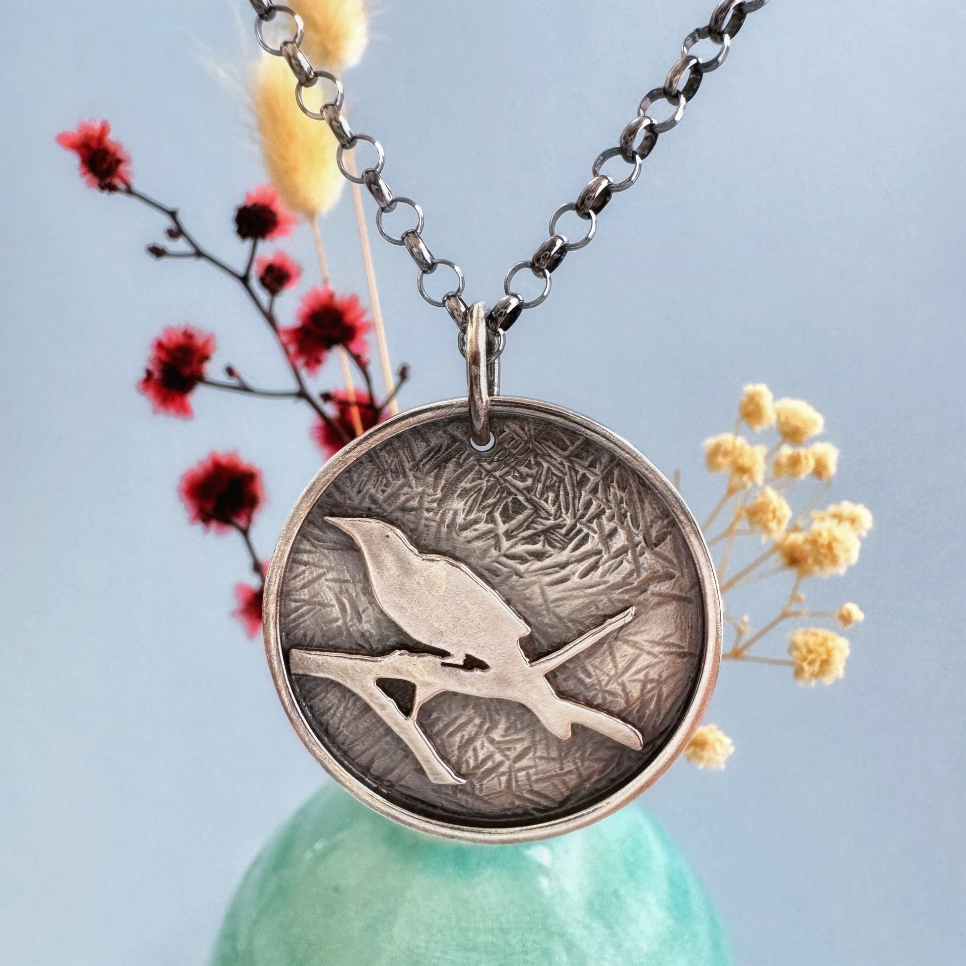 A round sterling silver pendant hangs from a rolo style chain. The pendant features a raised raven or crow sitting on the branch of a tree. The background has been hammered to represent leaves. The necklace is hanging in front of a pale green vase with cream and dark pink dried flowers, in front of a pale blue background.