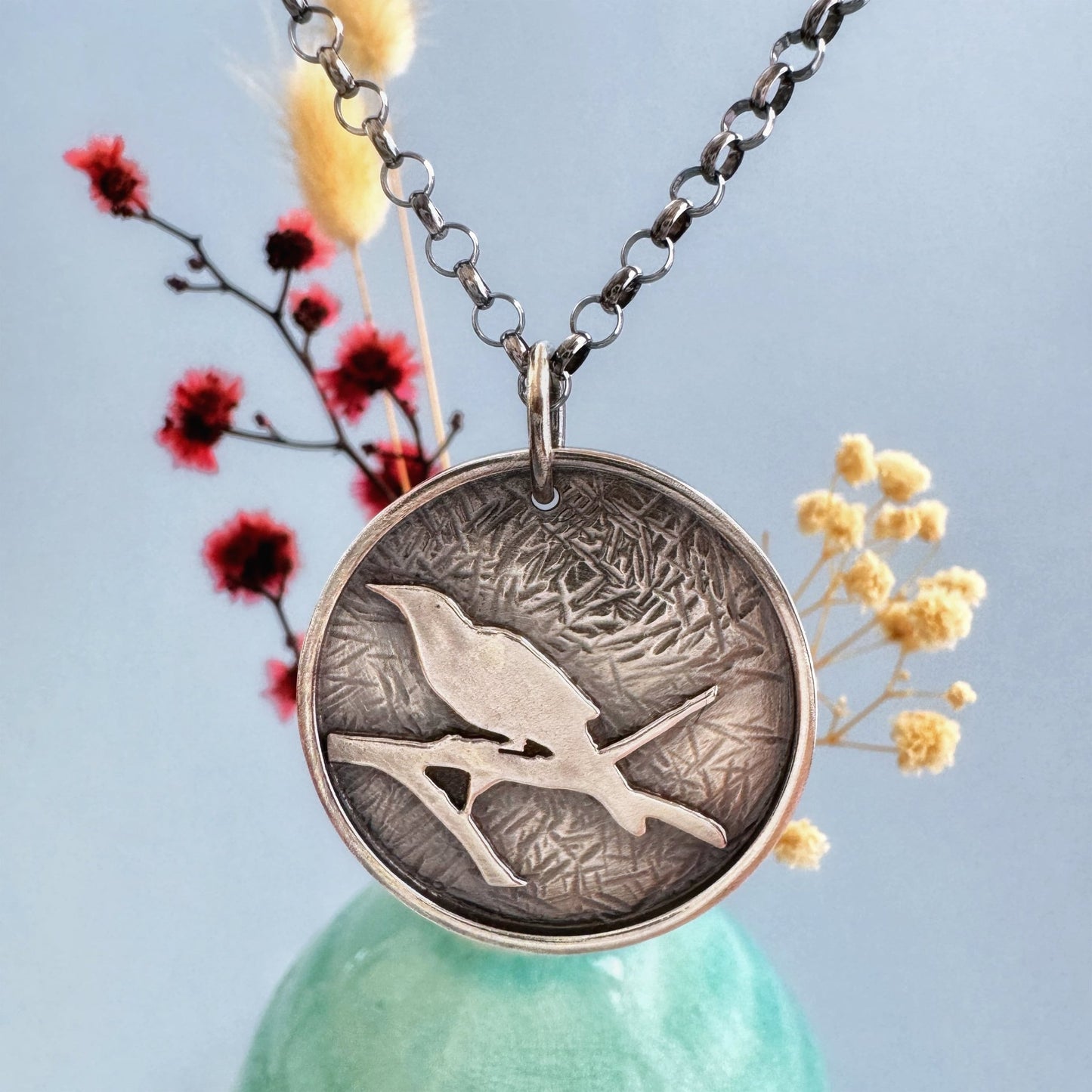 A round sterling silver pendant hangs from a rolo style chain. The pendant features a raised raven or crow sitting on the branch of a tree. The background has been hammered to represent leaves. The necklace is hanging in front of a pale green vase with cream and dark pink dried flowers, in front of a pale blue background.