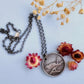 A round sterling silver pendant hangs from a rolo style chain. The pendant features a raised raven or crow sitting on the branch of a tree. The background has been hammered to represent leaves. The necklace is lying on a pale blue background, surrounded by cream and dark pink dried flowers.