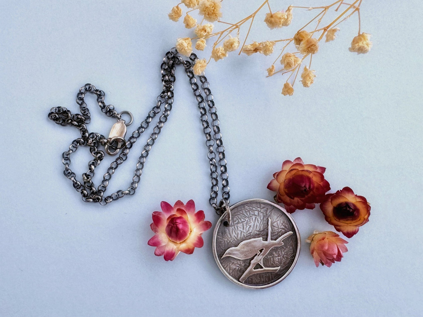 A round sterling silver pendant hangs from a rolo style chain. The pendant features a raised raven or crow sitting on the branch of a tree. The background has been hammered to represent leaves. The necklace is lying on a pale blue background, surrounded by cream and dark pink dried flowers.