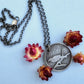 A round sterling silver pendant hangs from a rolo style chain. The pendant features a raised raven or crow sitting on the branch of a tree. The background has been hammered to represent leaves. The necklace is lying on a pale blue background, surrounded by cream and dark pink dried flowers.
