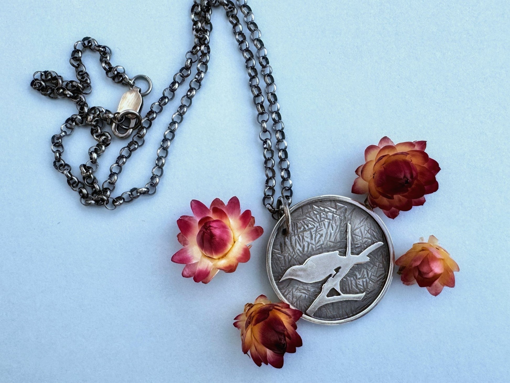 A round sterling silver pendant hangs from a rolo style chain. The pendant features a raised raven or crow sitting on the branch of a tree. The background has been hammered to represent leaves. The necklace is lying on a pale blue background, surrounded by cream and dark pink dried flowers.
