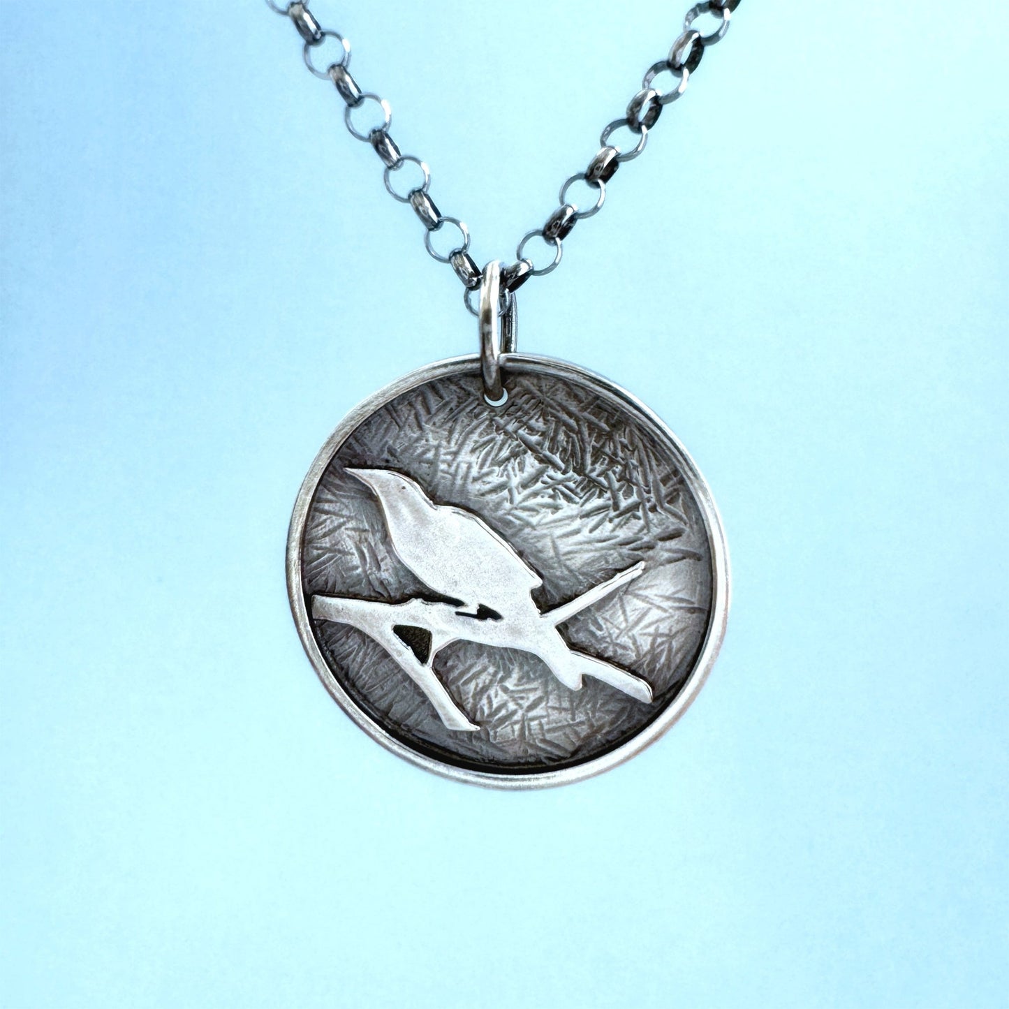 A round sterling silver pendant hangs from a rolo style chain. The pendant features a raised raven or crow sitting on the branch of a tree. The background has been hammered to represent leaves. The necklace is hanging in front of a pale blue background.