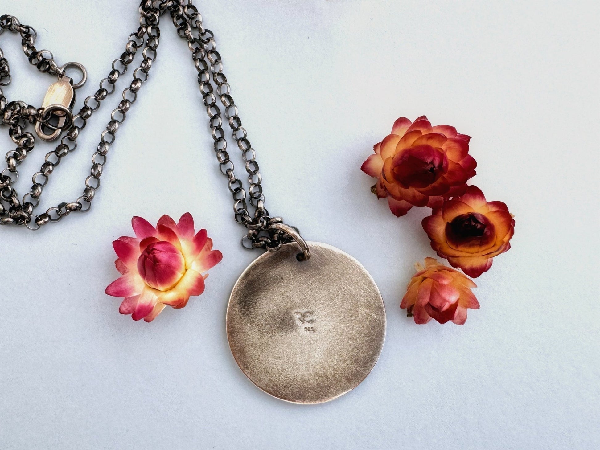 The back of the raven/crow pendant, showing our RC mark and the 925 quality stamp. The necklace is lying on a pale blue background, surrounded by cream and dark pink dried flowers.