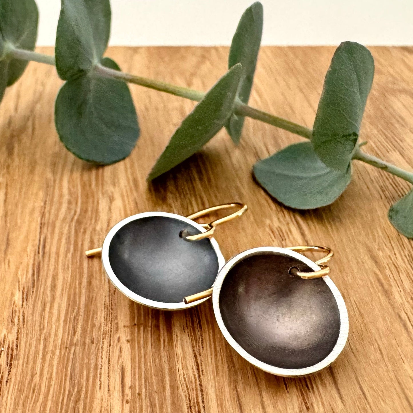 A pair of oxidised sterling silver disc dangle earrings with 14/20Y gold filled ear wires, on a wooden background with a small sprig of eucalyptus leaves. 