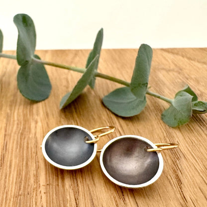 A pair of oxidised sterling silver disc dangle earrings with 14/20Y gold filled ear wires, on a wooden background with a small sprig of eucalyptus leaves. 