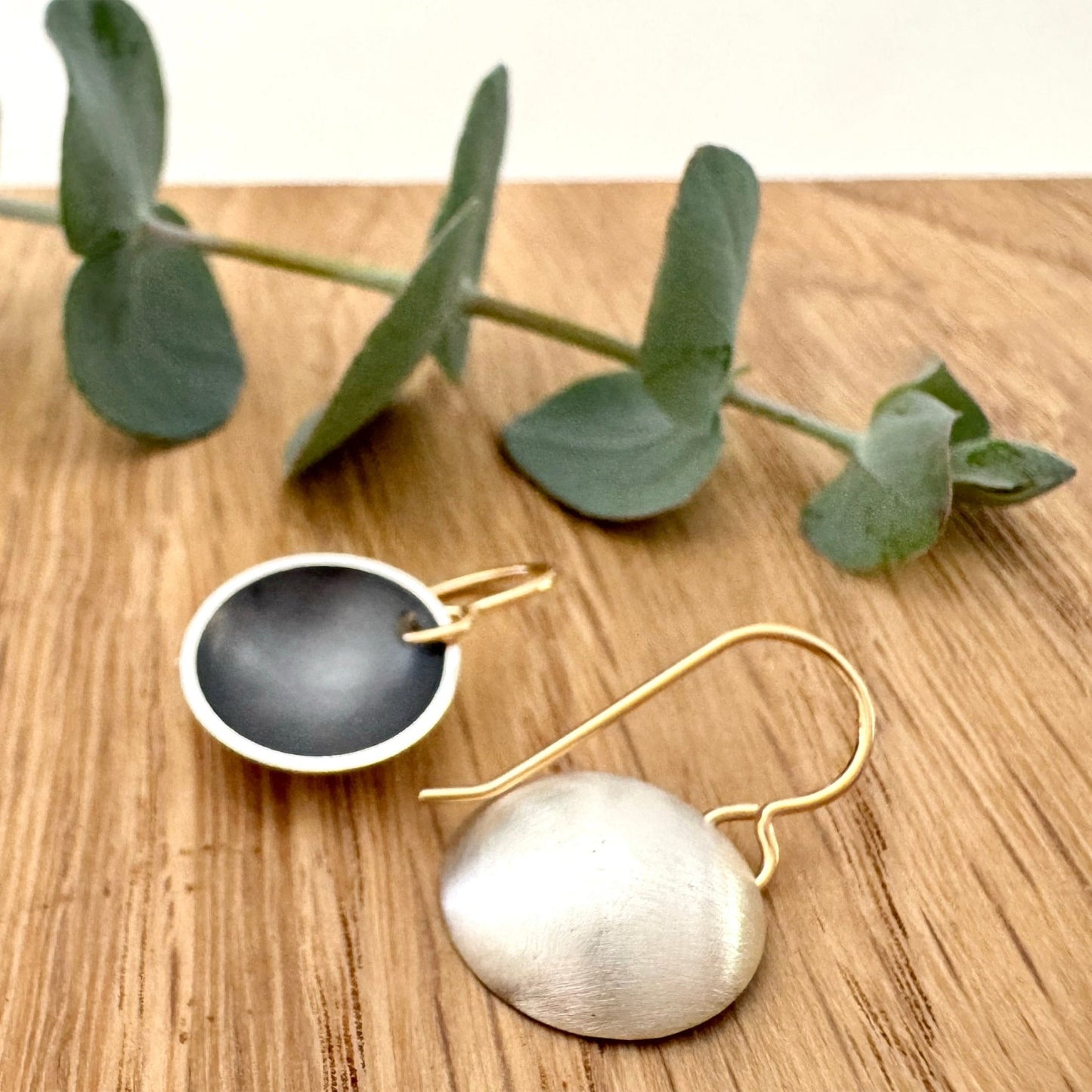 A pair of oxidised sterling silver disc dangle earrings with 14/20Y gold filled ear wires, on a wooden background with a small sprig of eucalyptus leaves. 
