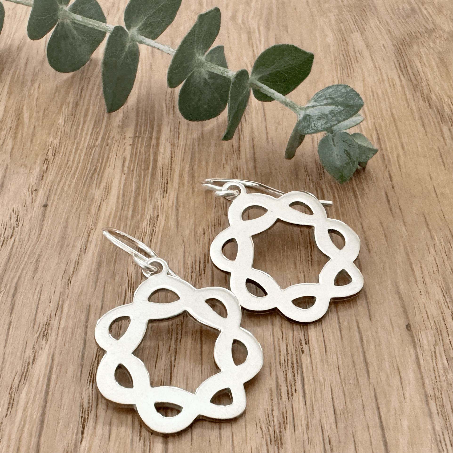 Sterling silver celtic knot hoop earrings on a wooden surface with a small sprig of eucalyptus.