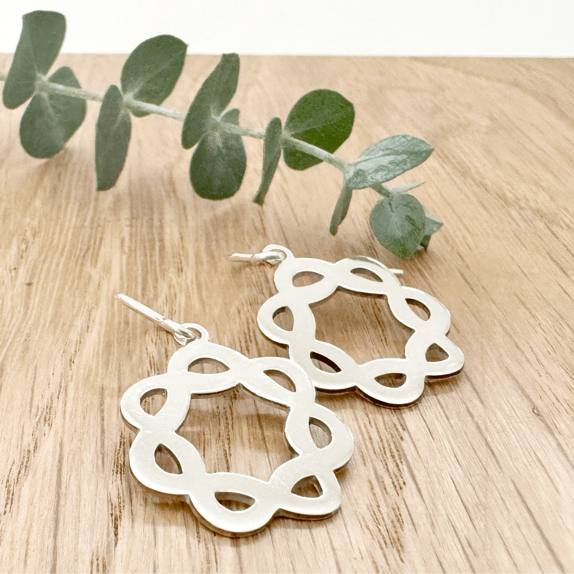 Sterling silver celtic knot hoop earrings on a wooden surface with a small sprig of eucalyptus.